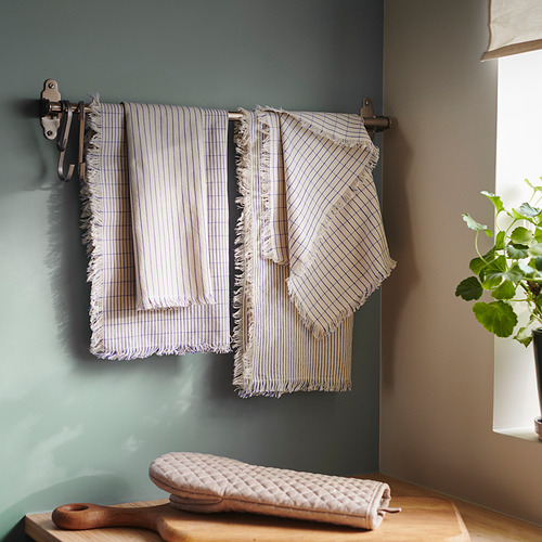 Toallas de rayas blancas con flecos cuelgan en perchero de pared, pared azul claro. Tabla de cortar de madera y guante de cocina cerca, planta verde en la ventana.