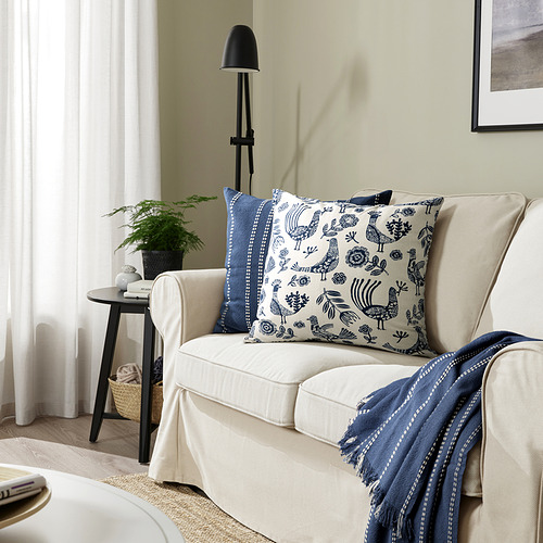 Almohada KRUTBRÄNNARE, beige y azul, algodón, cuadrado, gallo y diseño floral. Más pequeña que la silla, junto a una lámpara alta.