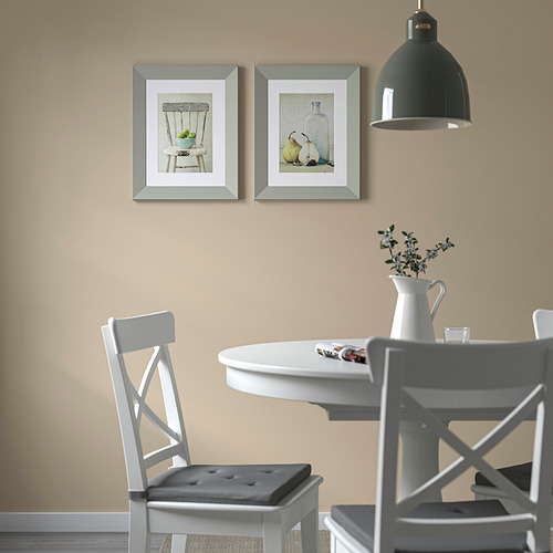 Dos cuadros enmarcados sobre mesa de comedor con sillas blancas, flores y luz colgante.