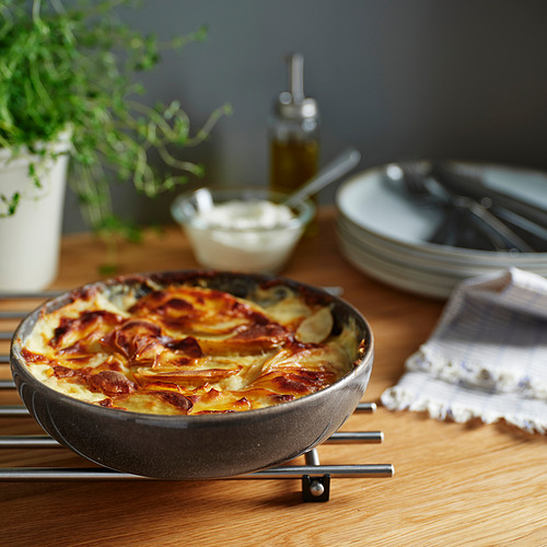 Plato de horno gris sobre salvamanteles con comida horneada, rodeado de platos, cubiertos y plantas.