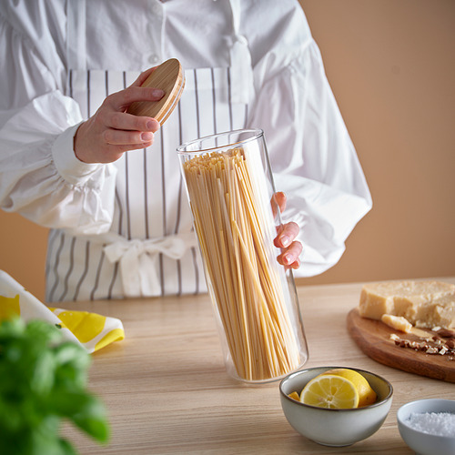 Una persona con delantal sostiene un frasco de espaguetis. La mesa tiene queso, limones y tabla de cortar.