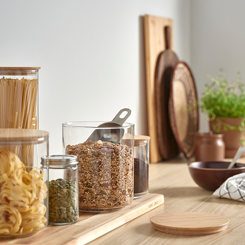 Encimera de cocina con frasco de granola, tapa de bambú, rodeada de frascos pequeños, tablas de madera y hierba.