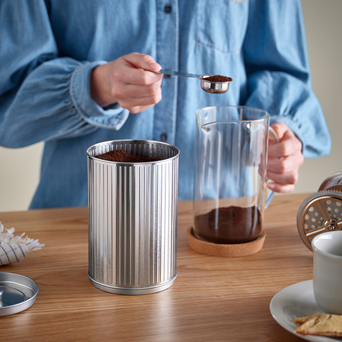 Una persona está colocando posos de café en un recipiente de metal sobre una mesa, con una prensa francesa de vidrio y una taza cerca.