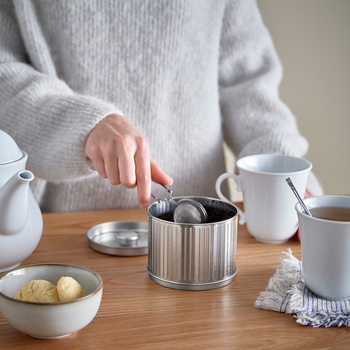 Persona preparando té en mesa de madera con colador, tetera, tazas y galletas.