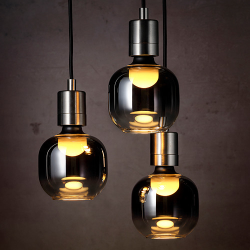 Cuelgan tres colgantes de vidrio gris ahumado, cada uno iluminado cálidamente, proyectando un brillo acogedor. La iluminación LED en el interior agrega elegancia moderna.