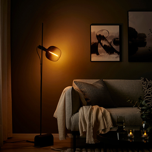 Acogedora sala de estar con lámpara de pie negra, luz cálida, sofá gris, almohadas, mantas y arte de pared. La lámpara es alta y delgada con una bombilla única.