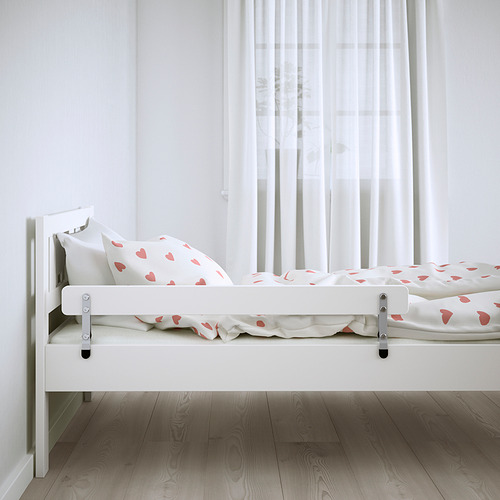 Pequeña cama infantil blanca con sábanas con estampado de corazones y barandillas junto a la ventana.