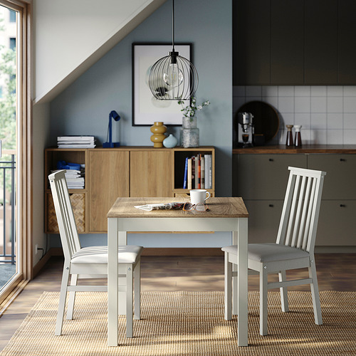 Moderna zona de comedor con mesa y sillas ÅLHULT, de madera clara y blanca, colocada en un espacio compacto con una cocina al fondo.