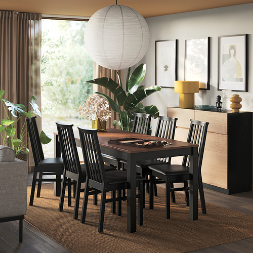 Una zona de comedor moderna con una silla oscura ÅLHULT colocada alrededor de una mesa de madera, que complementa la decoración contemporánea circundante y los grandes ventanales.