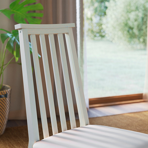 Acogedora escena interior: silla de listones blancos junto a la ventana, planta exuberante, cortina marrón, suelo de madera clara.