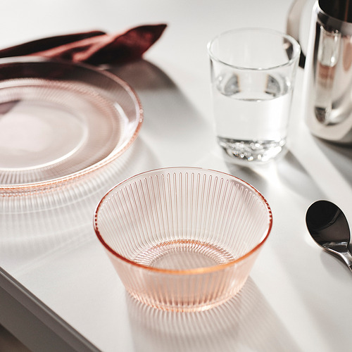 Un cuenco de cristal HAVSGÄDDA teñido de rosa y textura acanalada, pequeño y delicado, colocado sobre un mantel blanco junto a un vaso de agua clara y un plato.