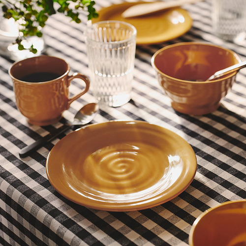 Vajilla SANDSKÄDDA en amarillo ocre, incluyendo platos, tazas y un vaso. Gres de tacto rústico, apilado sobre mantel de cuadros.