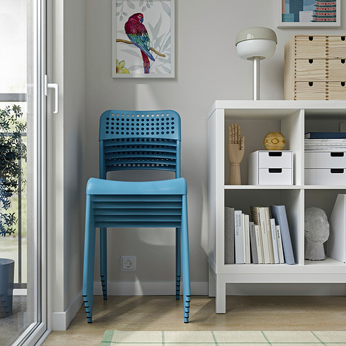 Silla de plástico azul contra una pared, junto a una estantería con libros, artículos de decoración y una lámpara.