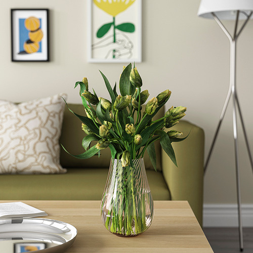 Jarrón de vidrio PÅSKÖTRÄD con remolinos que sostiene flores de color amarillo verdoso sobre una mesa de madera junto a la lámpara y el sofá.