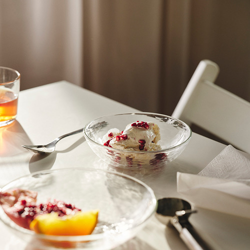 El tazón de vidrio con diseño martillado contiene helado cubierto con semillas de granada, con vaso, tenedor y cuchara vacíos sobre una mesa blanca.