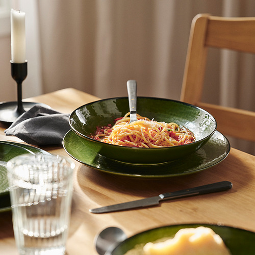 Gran cuenco verde de espaguetis sobre mesa de madera con tenedor, cuchillo, vaso, vela y servilleta.