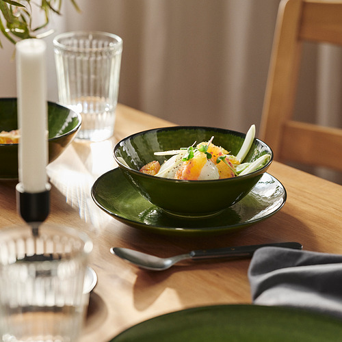 Una mesa de madera con un cuenco de comida de color verde oscuro, un plato, un vaso, una cuchara y una vela.