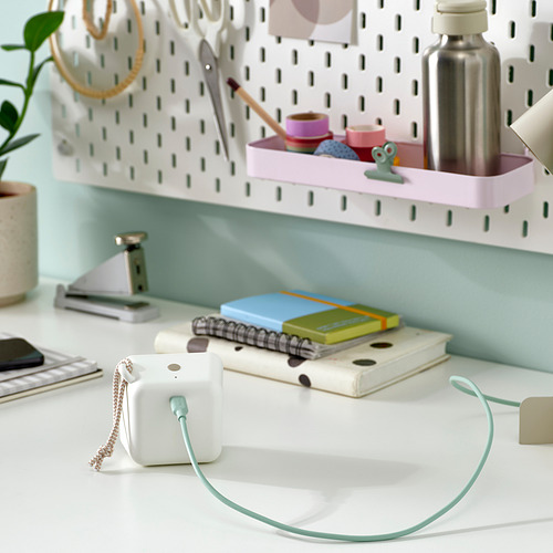 Un escritorio blanco con un cable usb-c verde conectado a un pequeño dispositivo, notebooks y una planta. Un tablero perforado con material de oficina al fondo.