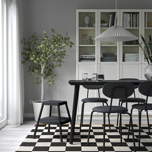 Comedor moderno con taburetes negros, sillas, mesa de madera, alfombra, lámpara, estanterías con libros y una planta en maceta.