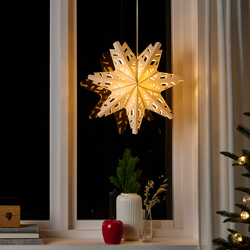 Lámpara STRÅLA, blanca, de papel, en forma de estrella, colgante, iluminada, colocada cerca de una ventana y un árbol de navidad, rodeada de decoraciones más pequeñas.