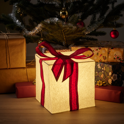 Pequeño cubo de luz brillante, de color crema con cinta roja, debajo de un árbol de Navidad, rodeado de regalos envueltos.