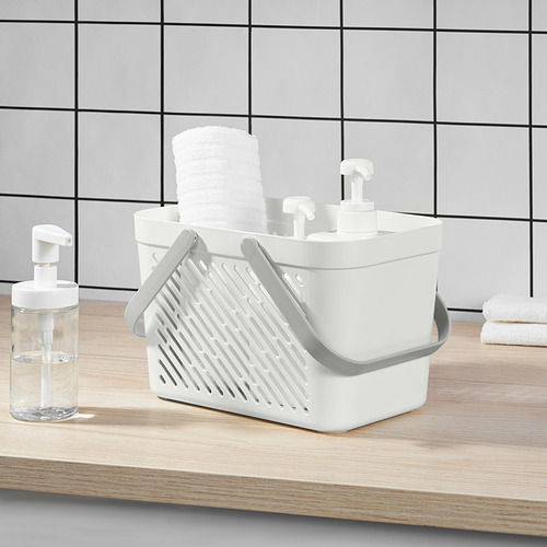 Cesta de ducha de plástico blanco con asas plegables, apilada con toallas blancas enrolladas, colocada sobre encimera de madera clara junto al dispensador de jabón transparente.