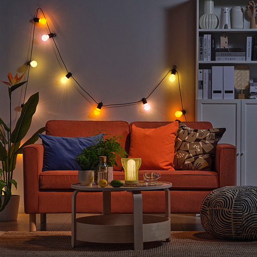 Acogedora sala de estar con sofá naranja, guirnaldas de luces, almohadas, mesa de café, plantas y estantería.