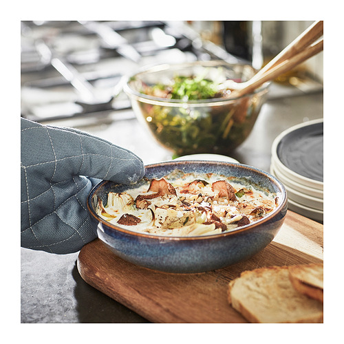 Una mano agarra un plato de cerámica vidriada azul lleno de comida horneada, junto a una ensaladera y pan sobre una tabla de madera.
