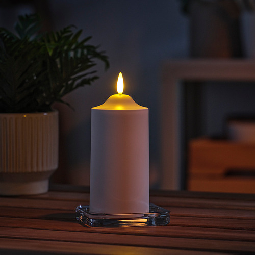 Vela LED blanca SMÅLOM de Ikea en soporte de vidrio, que brilla cálidamente sobre una mesa de madera junto a una planta verde.