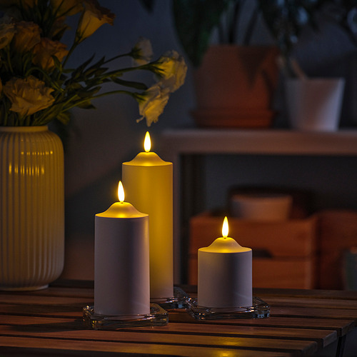 Tres velas SMÅLOM encendidas en blanco sobre una mesa de madera. Uno es más grande, dos son más pequeños. Acompañado de un jarrón amarillo con flores a la izquierda.