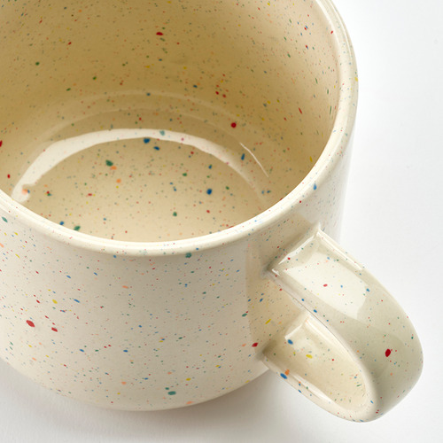 Taza de gres moteado con asa, base blanquecina, motas multicolores, textura suave, diseño sencillo.