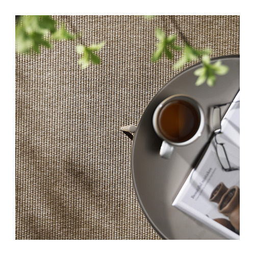 Una alfombra LÄTTBETONG beige y marrón claro para exteriores. Encima hay una taza de café. La alfombra tiene una superficie plana y texturizada y está bordeada de vegetación.