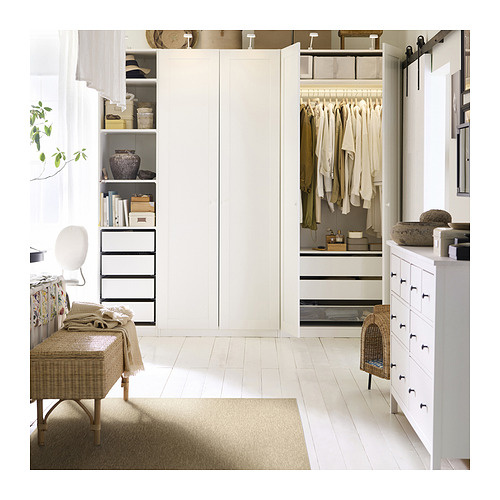 Dormitorio blanco moderno con alfombra beige, banco de mimbre y armario abierto con ropa, estantes y cajones.