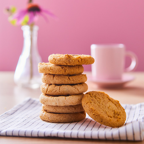 Una pila de galletas KAFFEREP de color marrón dorado sobre una tela a rayas, con una taza blanca y un jarrón con flores en el fondo.