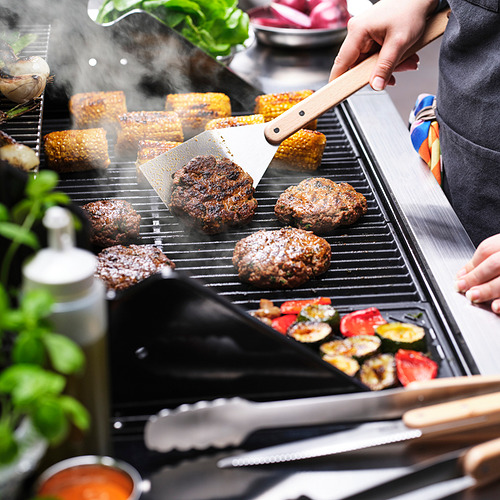 Persona cocinando hamburguesas y verduras en una parrilla con una espátula.