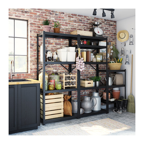 Estantes de almacenamiento de metal negro contra una pared de ladrillo, que sostienen varios artículos de cocina como frascos, ollas y utensilios, con decoración rústica.