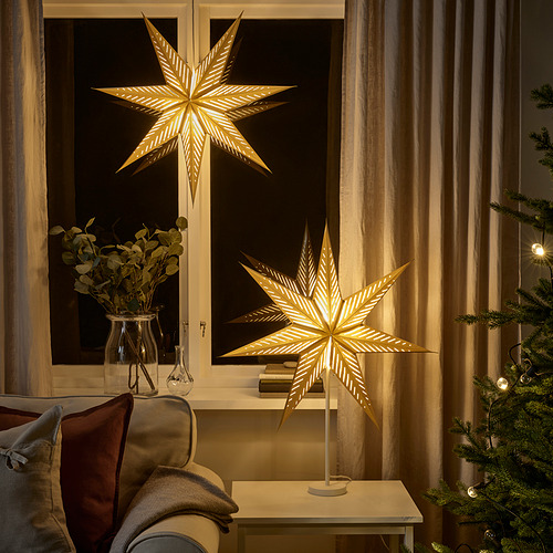 Gran lámpara dorada en forma de estrella, parte de la colección STRÅLA, se encuentra en la mesa junto al árbol de Navidad y el sofá. Resplandores suaves, iluminados.