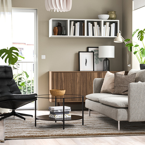 Sala de estar moderna con alfombra BANKKONTOR, sofá gris, mesa de centro, silla negra, almacenamiento de madera y plantas. El tamaño de las alfombras complementa los muebles.