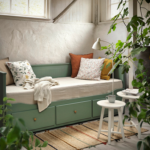 Sofá cama HEMNES en gris verdoso con ropa de cama color crema, taza y lámpara en la mesa auxiliar, alfombra a rayas. Moderno y acogedor.