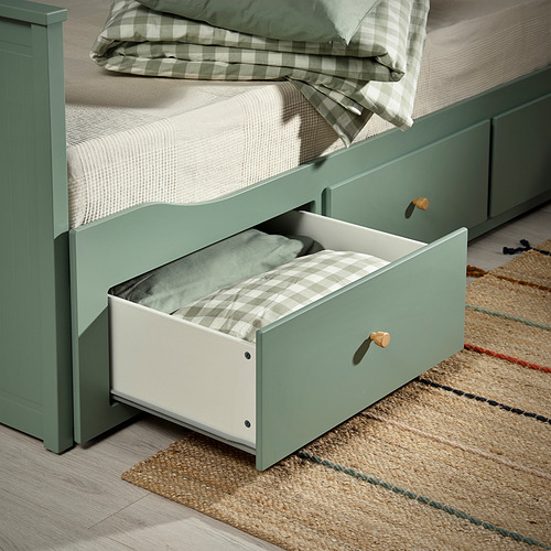 Sofá cama verde con sábanas a cuadros, estructura de madera, alfombra tejida.