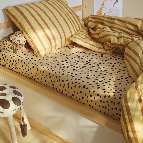 Cama infantil de temática safari con sábana bajera SANDLÖPARE en manchas marrones. Incluye almohadas a rayas, un peluche de leopardo y un taburete de jirafa cerca.