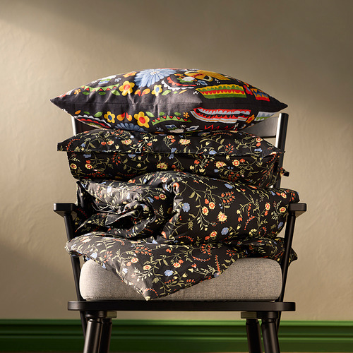 Almohadas BRUDKRUSBÄR: cojines florales negros apilados en la silla, más grandes abajo, más pequeños encima, diseños de flores vibrantes.