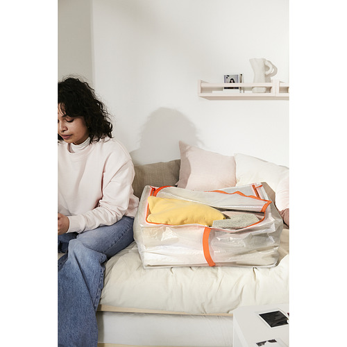 Persona sentada en la cama con un gran estuche de almacenamiento PÄRKLA, que contiene artículos amarillos doblados y pliegues grises. Pared blanca y estante en el fondo.