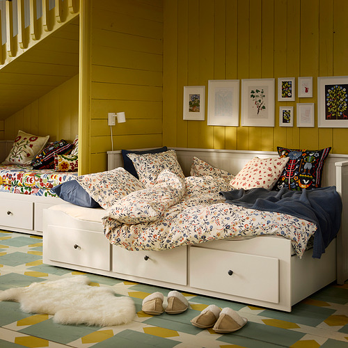 Habitación de color amarillo brillante con una cama BRUDKRUSBÄR blanca, ropa de cama floral, almohadas de colores, zapatillas y arte en la pared.