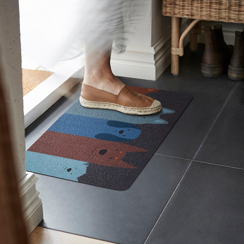 Persona pisando la esterilla TURISTVÄG . Colores marrón y azul, parte superior de poliéster reciclado, parte trasera de goma. Forma grande y rectangular.