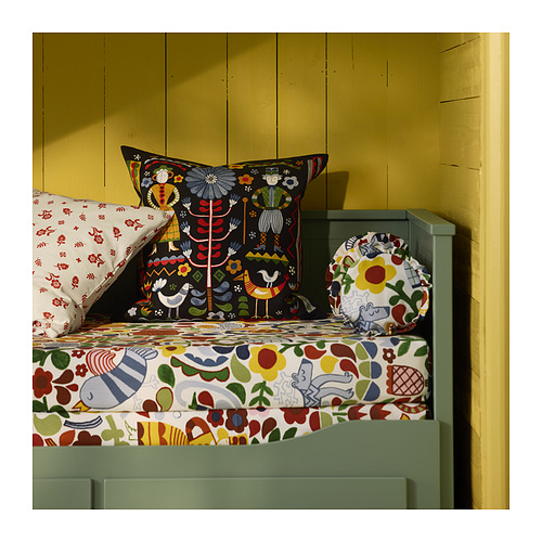 Cama con almohadas y mantas coloridas y estampadas, con diseños de arte popular, frente a una pared amarilla.