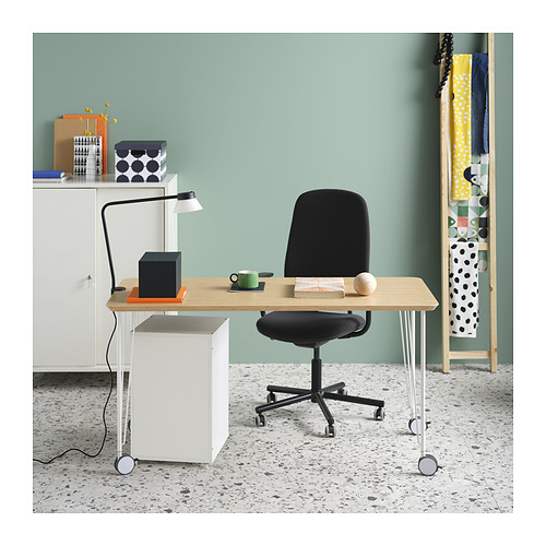 Moderna oficina en casa con escritorio de madera clara, silla negra y gabinete blanco. Pared verde, lámpara y elementos decorativos.