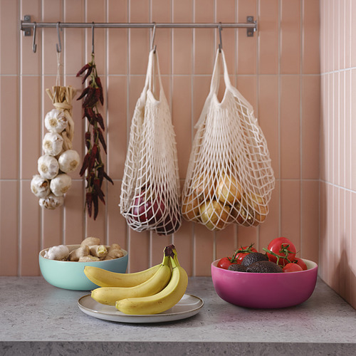 Encimera de cocina con productos frescos. Dos cuencos de colores: verde azulado, magenta y plateado. Bolsas de red con cebollas. Pared de azulejos color melocotón.