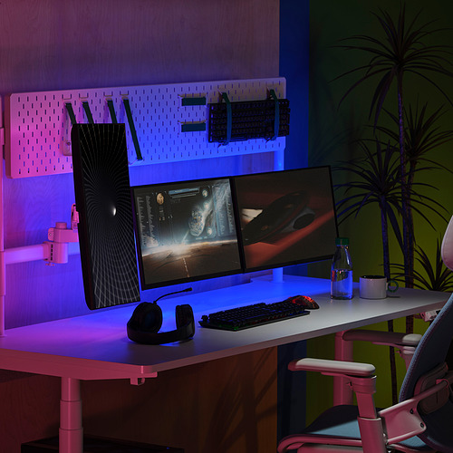 Escritorio moderno con monitores duales, teclado, mouse, auriculares para juegos y luces led. Silla de oficina rosa, planta araña, espacio de trabajo elegante.