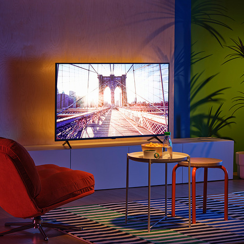 Moderno salón con tv de pantalla plana que muestra un puente. Silla roja brillante, pequeñas mesas redondas y alfombra colorida con plantas.